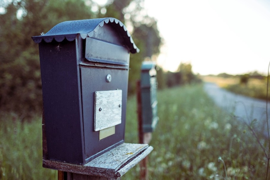 Ein Briefkasten an einer Straße. Im Hintergrund sind verschwommen Büsche, Wiese und Bäume zu erkennen.